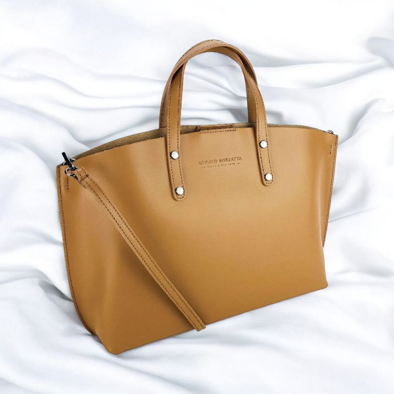 A tan leather tote bag rests elegantly on a pristine white bed, showcasing its stylish design and color contrast.