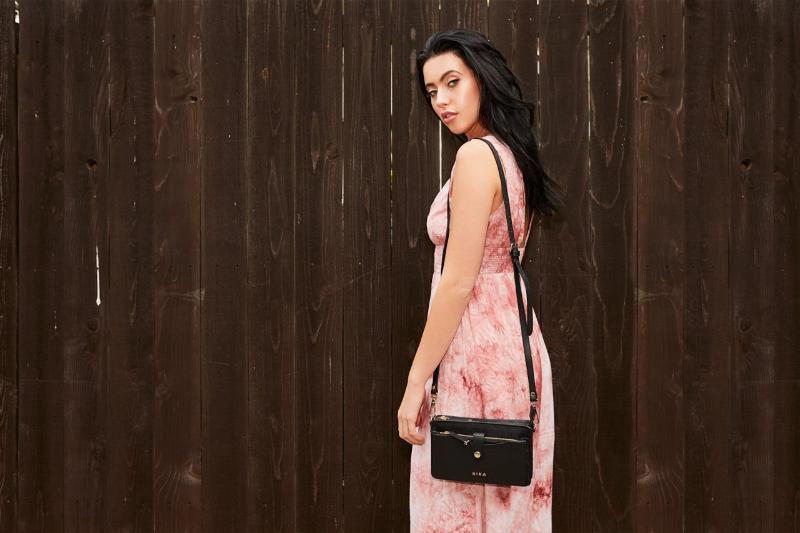 A woman in a lovely pink dress stands next to a wooden fence with a black crossbody bag on her shoulder.