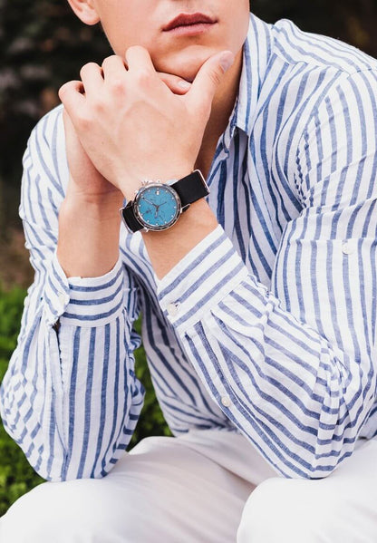 A man in a blue and white striped shirt and white pants, showcasing stylish ice-blue watches on his wrists.