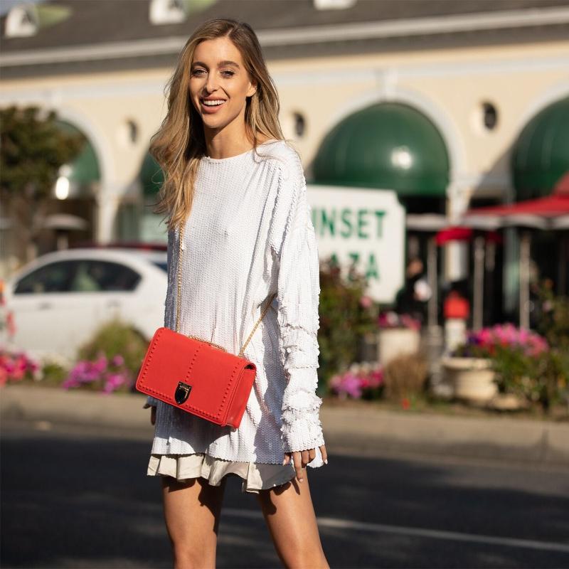 A woman in a white shirt and skirt strolls down the street, carrying a vibrant red purse.